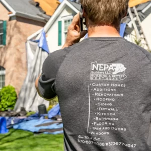 Man talking on cell phone looking at a roof replacement with his back to the camera. On the back of the T-shirt is NEPA Builders LLC Building a Better Northeast.