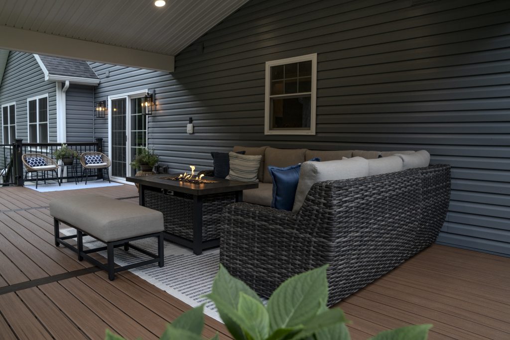 Cozy corner of a covered porch addition featuring a wicker sectional sofa with blue pillows and a rectangular fire pit table.