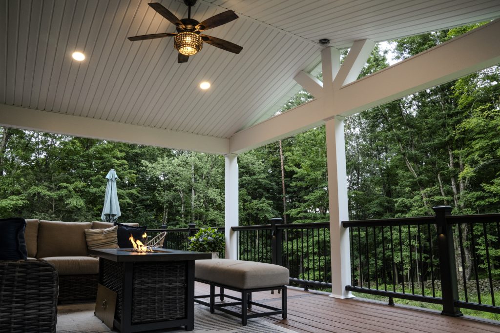 Cozy outdoor seating area on a composite deck with a sectional sofa and ottoman, located under a new roof extension attached to a gray vinyl-sided home.
