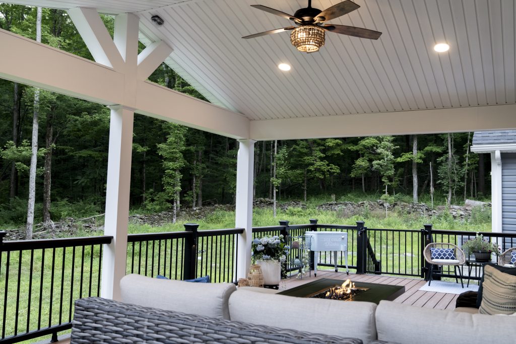 Interior view of a custom covered porch addition in Clark's Summit, PA, featuring a white beadboard ceiling, ceiling fan, and a wicker sectional arranged around a fire pit table.