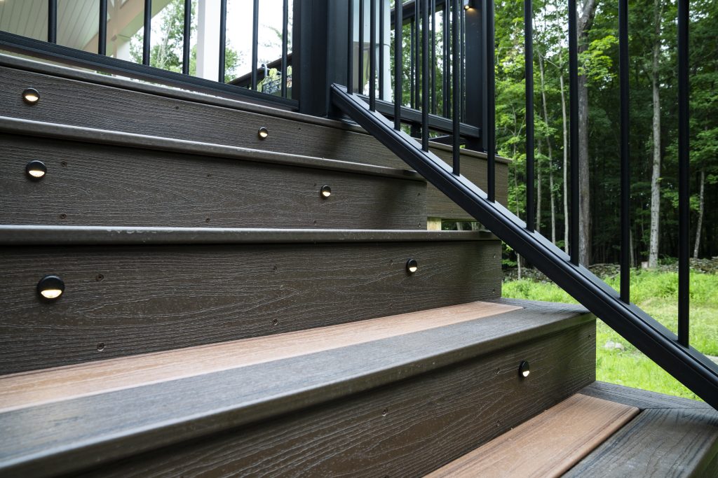 Close-up detail of composite deck stairs featuring built-in low-voltage riser lights and a black aluminum handrail for safety and curb appeal.
