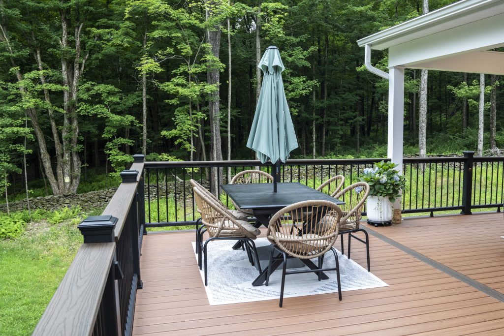 Open deck dining space featuring a table with a sage green umbrella, framed by white porch posts and a wooded Pennsylvania backyard.