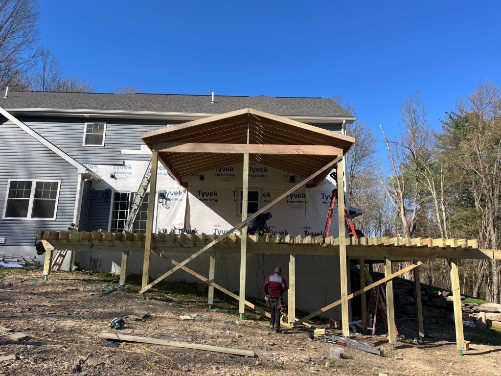 Framing of a new gable roof extension over a wooden deck, showing exposed rafters, Tyvek house wrap, and structural posts during a renovation project.