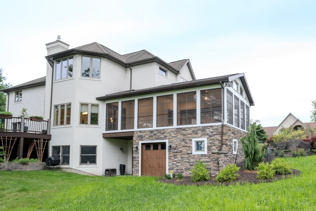 Rear view of a custom Eze-Breeze enclosed outdoor living addition featuring stone masonry, expansive windows, and a new garage in Dalton, PA.