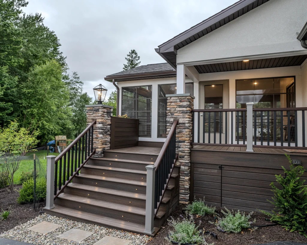 TimberTech deck stairs with integrated LED lighting lead to a custom three-season outdoor living space featuring cultured stone columns and an Eze-Breeze enclosure in Dalton, PA.